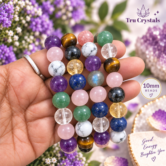 A hand holds three beaded stretch bracelets made of round 10mm crystals from Tru Crystals. The bracelets feature a variety of colorful gemstone beads including purple, green, pink, blue, yellow, brown, and clear quartz.