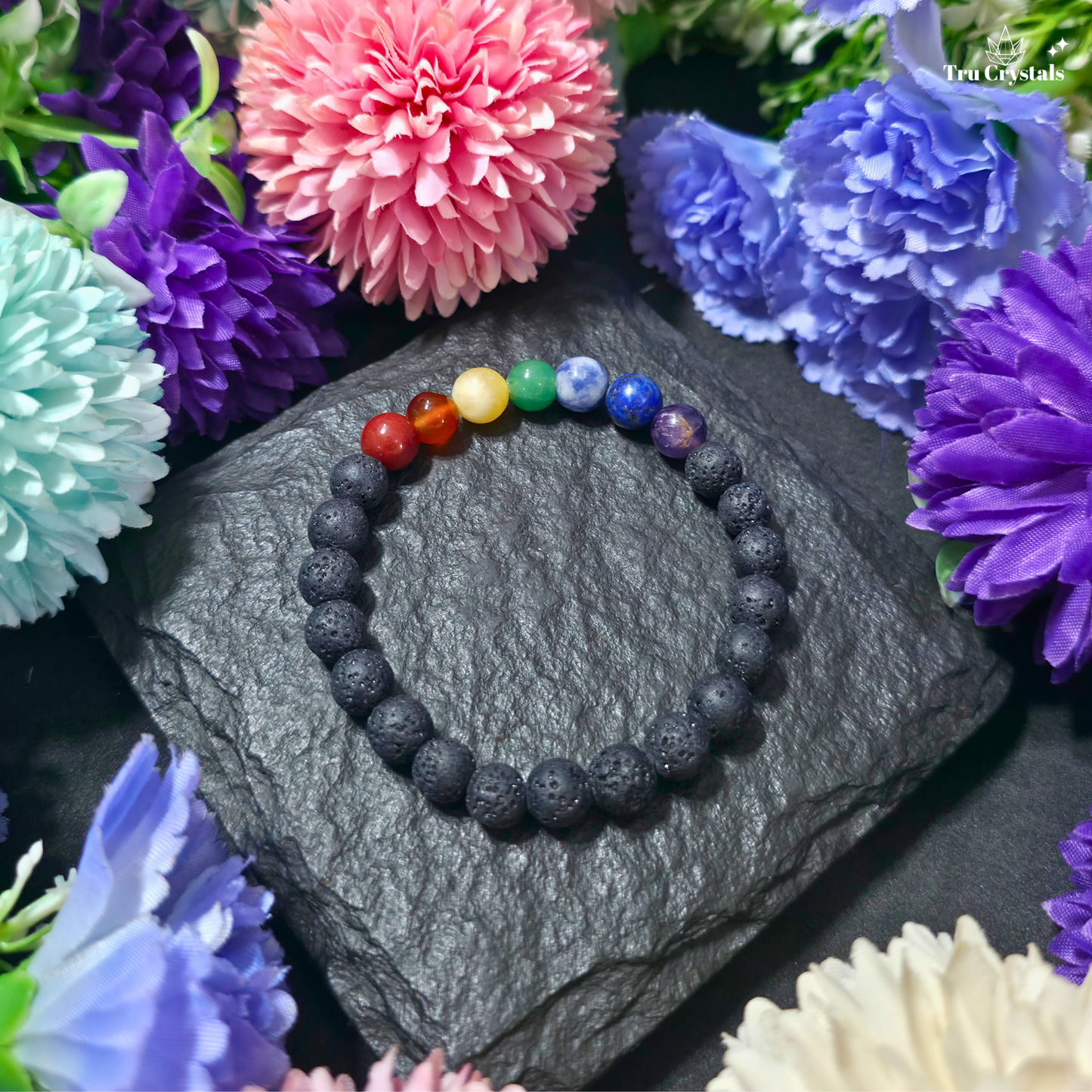 A beaded stretch bracelet with seven colorful chakra beads (red, orange, yellow, green, blue, purple, and dark blue) and matte black lava stone beads, displayed on a dark slate stone surrounded by artificial flowers.