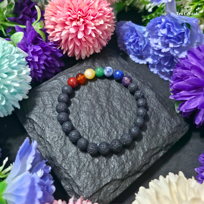 A beaded stretch bracelet with seven colorful chakra beads (red, orange, yellow, green, blue, purple, and dark blue) and matte black lava stone beads, displayed on a dark slate stone surrounded by artificial flowers.