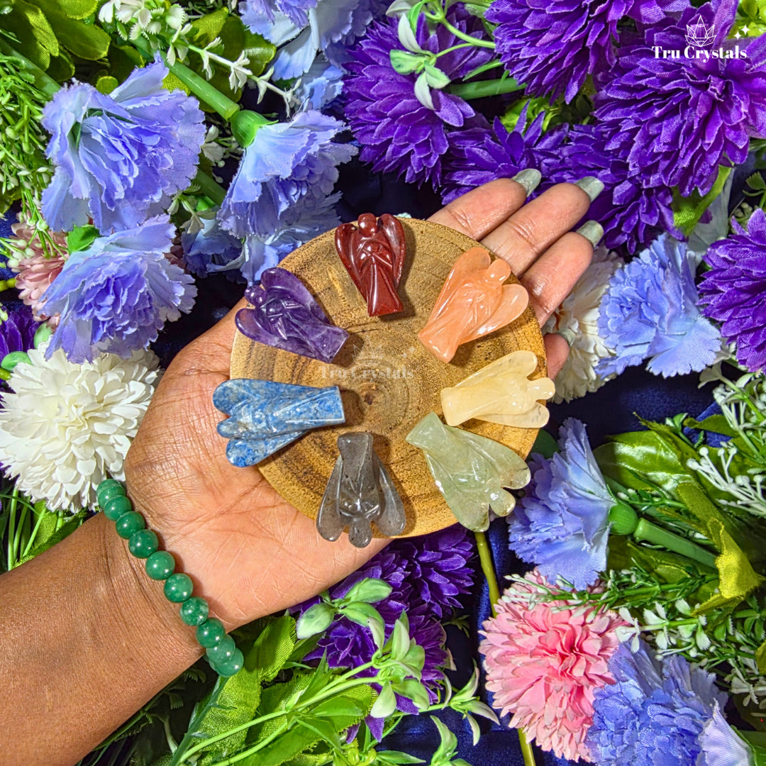 A hand holds a round wooden tray displaying seven carved crystal angels in different colors, surrounded by purple, blue, pink, and white flowers. A green beaded bracelet is worn on the wrist, and a 'Trucrystal' logo is visible in the top right corner.