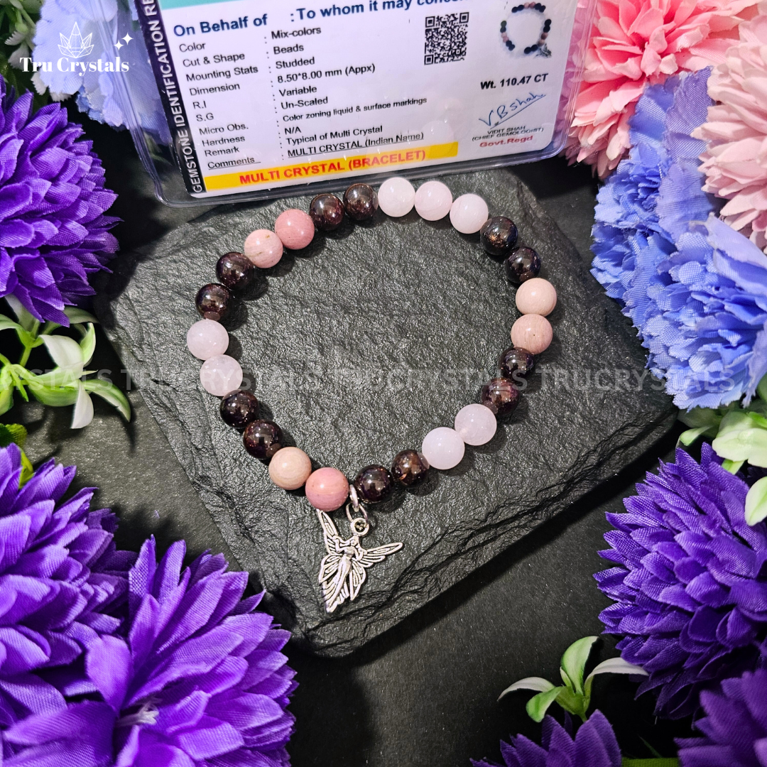 A multicolored stone bracelet displayed on a dark stone surface surrounded by various colorful flowers, with a small heart-shaped pendant hanging from the bracelet. The bracelet contains rose quartz , Rhodonite , rhodochrosite & garnet .