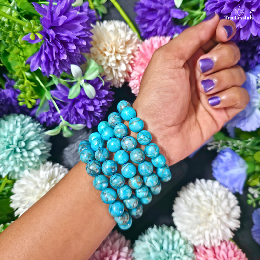 A close-up of a person's wrist wearing a African blue Turquoise beaded bracelet, with purple nail polish and a background of colorful artificial flowers.