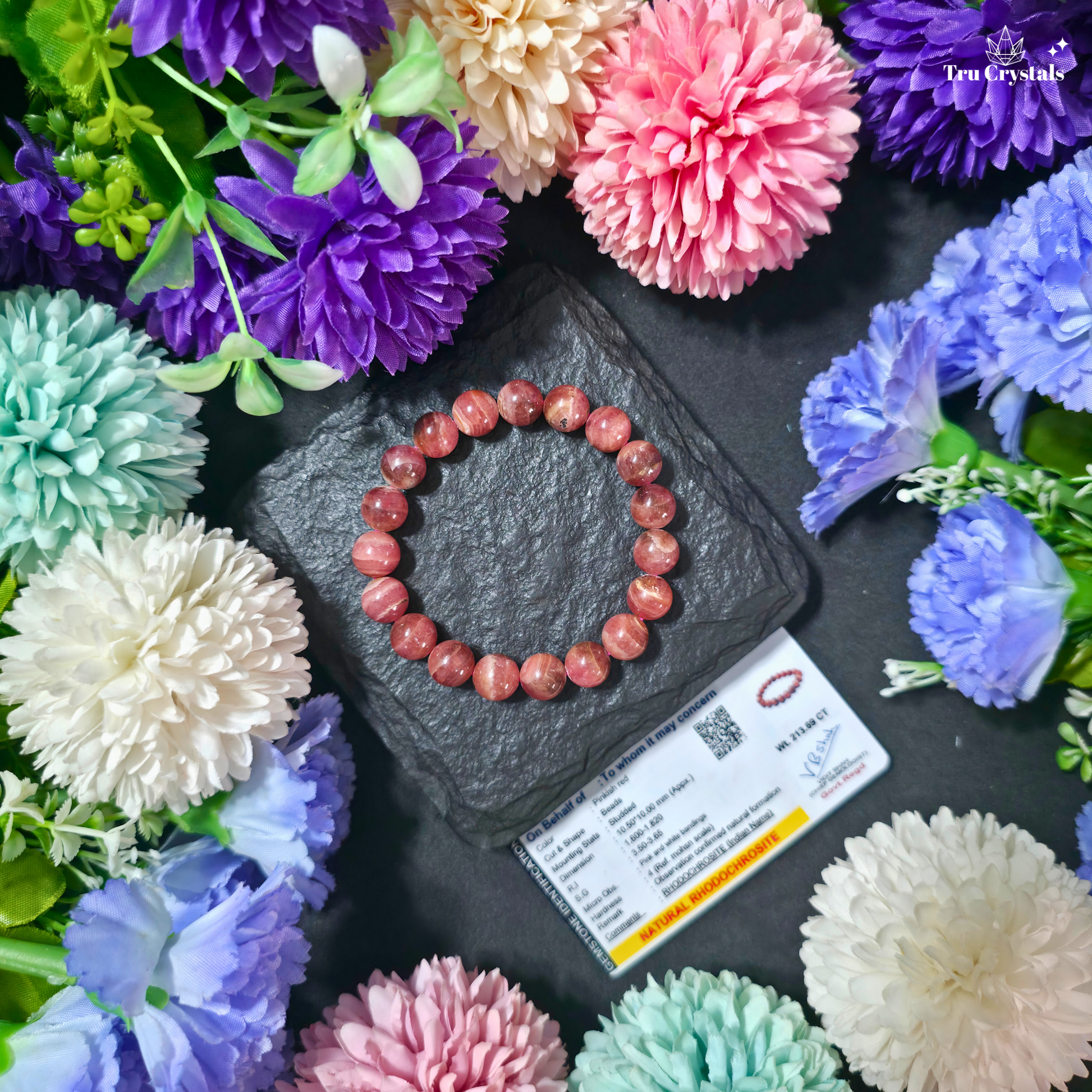 A Rhodochrosite bracelet displayed on a dark stone surface with vibrant pink flowers around it. A Gemological Certificate is also visible in the background, indicating the bracelet's authenticity and quality.