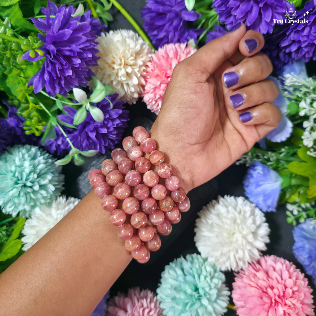 A person's hand and wrist wearing a multi-strand beaded bracelet made of pinkish-brown polished round beads, set against a background of colorful artificial flowers.