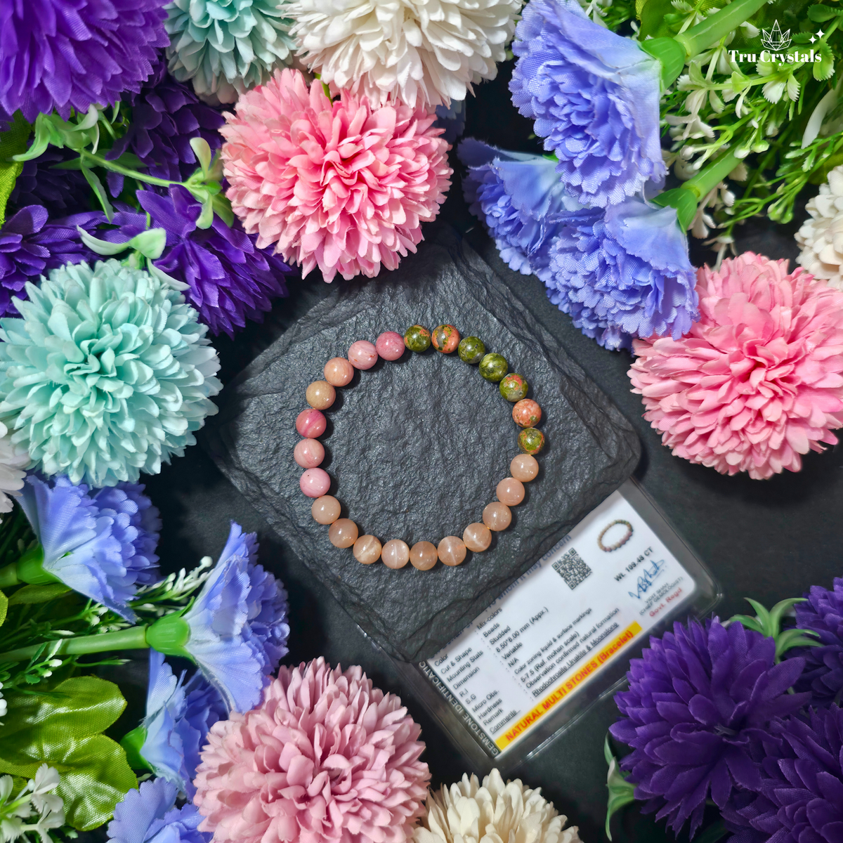A beaded bracelet made of pink, peach, and green marbled beads is arranged in a circle on a dark slate block, surrounded by colorful artificial flowers and a small product card. The brand name 'Trucrystals' is visible in the top right corner.