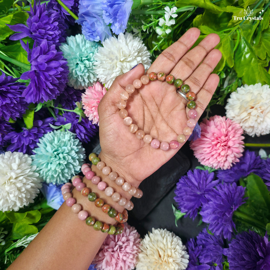 A multicolored gemstone bracelet containing moonstone, rhodochrosite and unakite displayed on a wooden surface with flowers and a colorful container in the background.