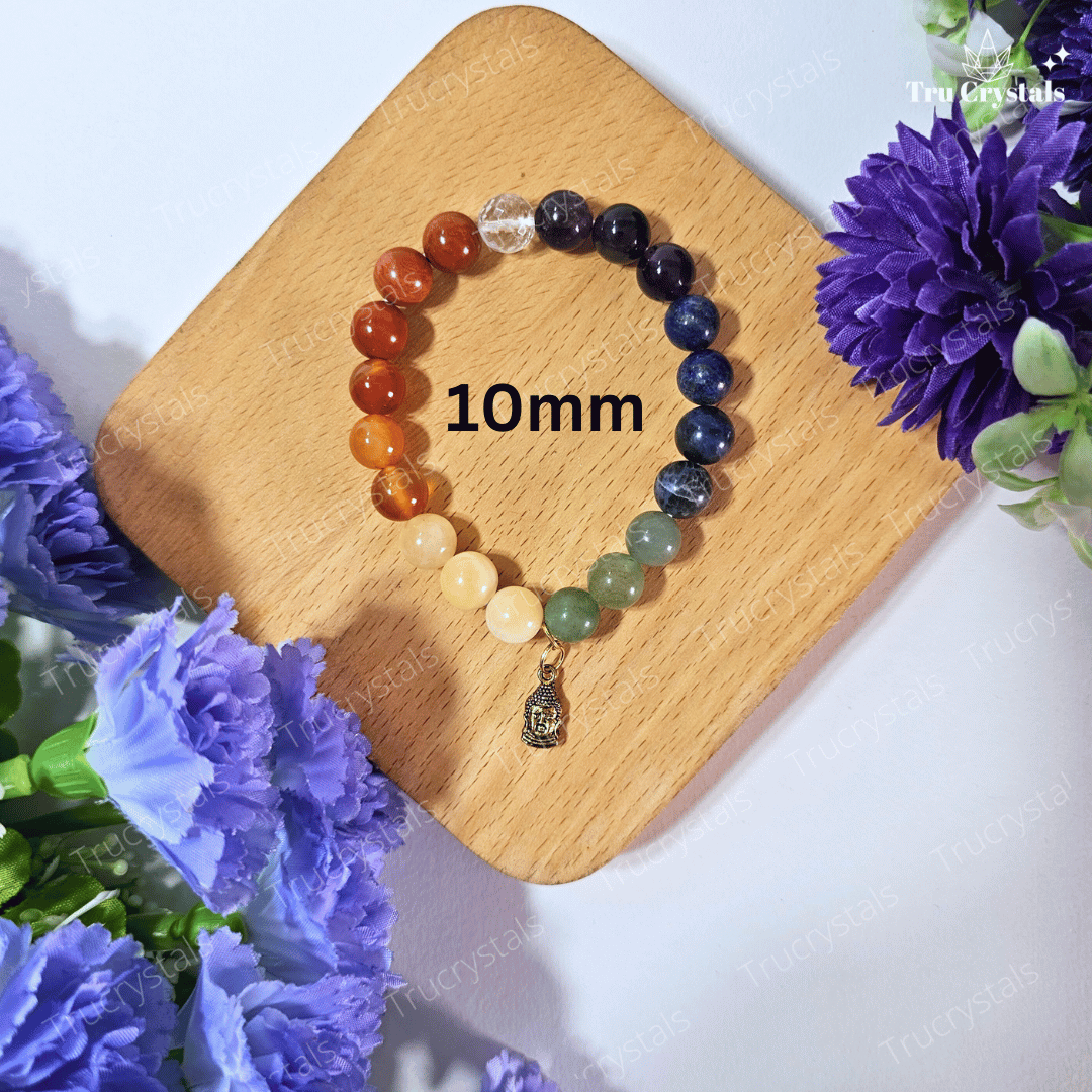 A multicolored 7 chakra bracelet with a Buddha charm, displayed on a wooden surface with a background of purple flowers and a clear quartz crystal.