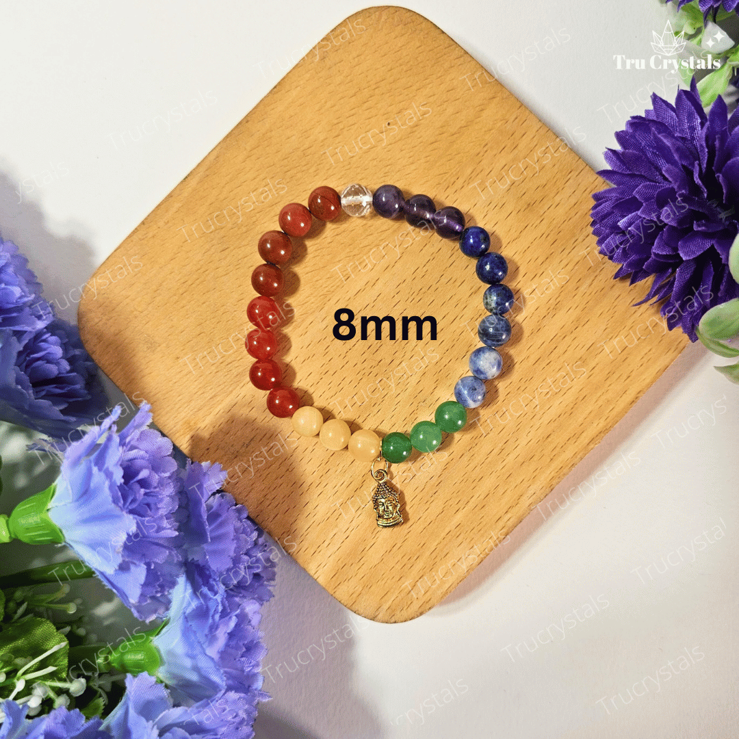 A multicolored 7 chakra bracelet with a Buddha charm, displayed on a wooden surface with a background of purple flowers and a clear quartz crystal.