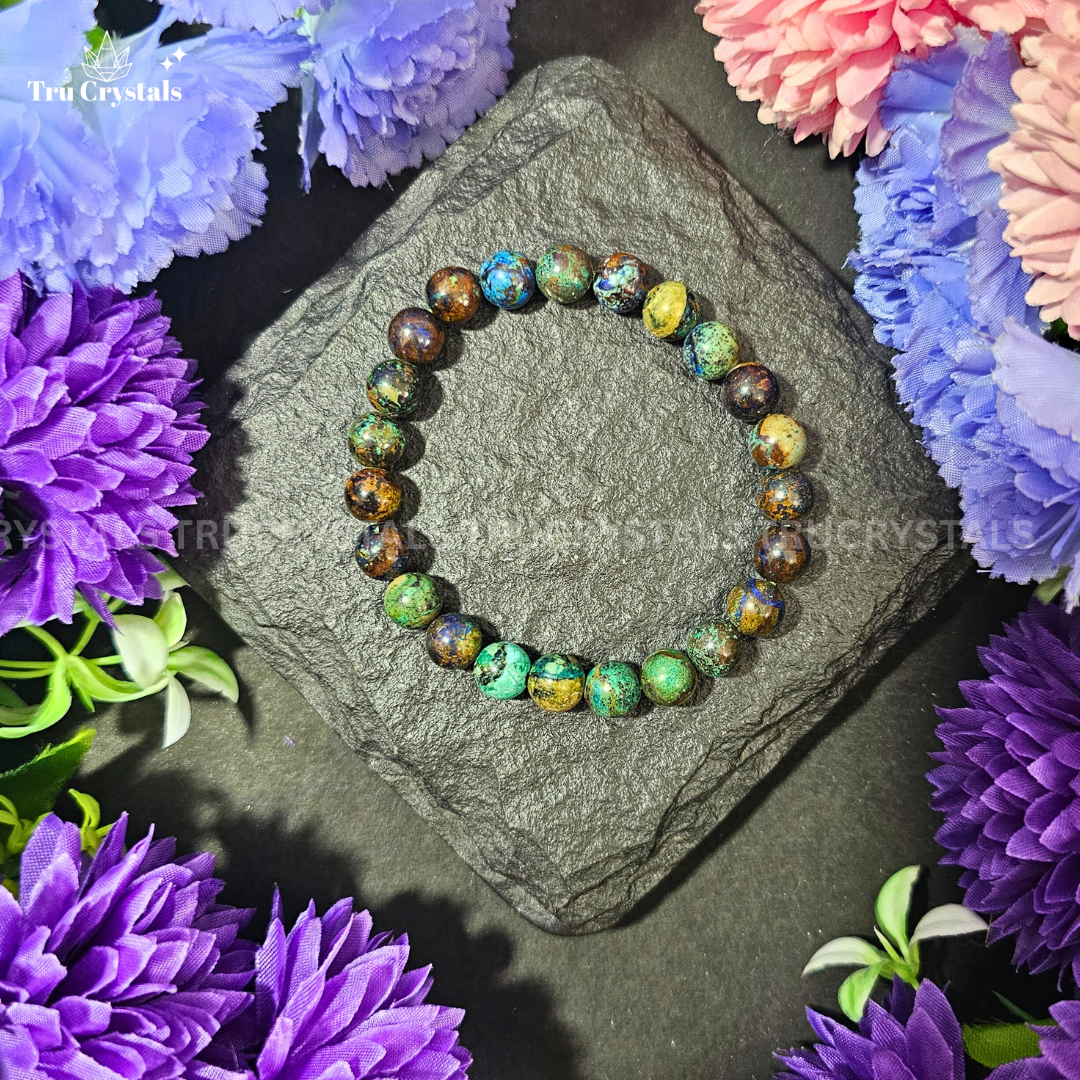 A Chrysocolla bracelet displayed on a rock, with a colorful gemstone certified by a laboratory, and flowers in the background.