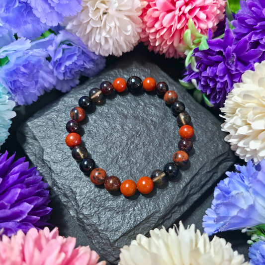 A bracelet made with beads of Red Jasper, Garnet, Smoky Quartz, Black Tourmaline, and Mahogany Obsidian, displayed on a dark stone surface surrounded by colorful flowers.