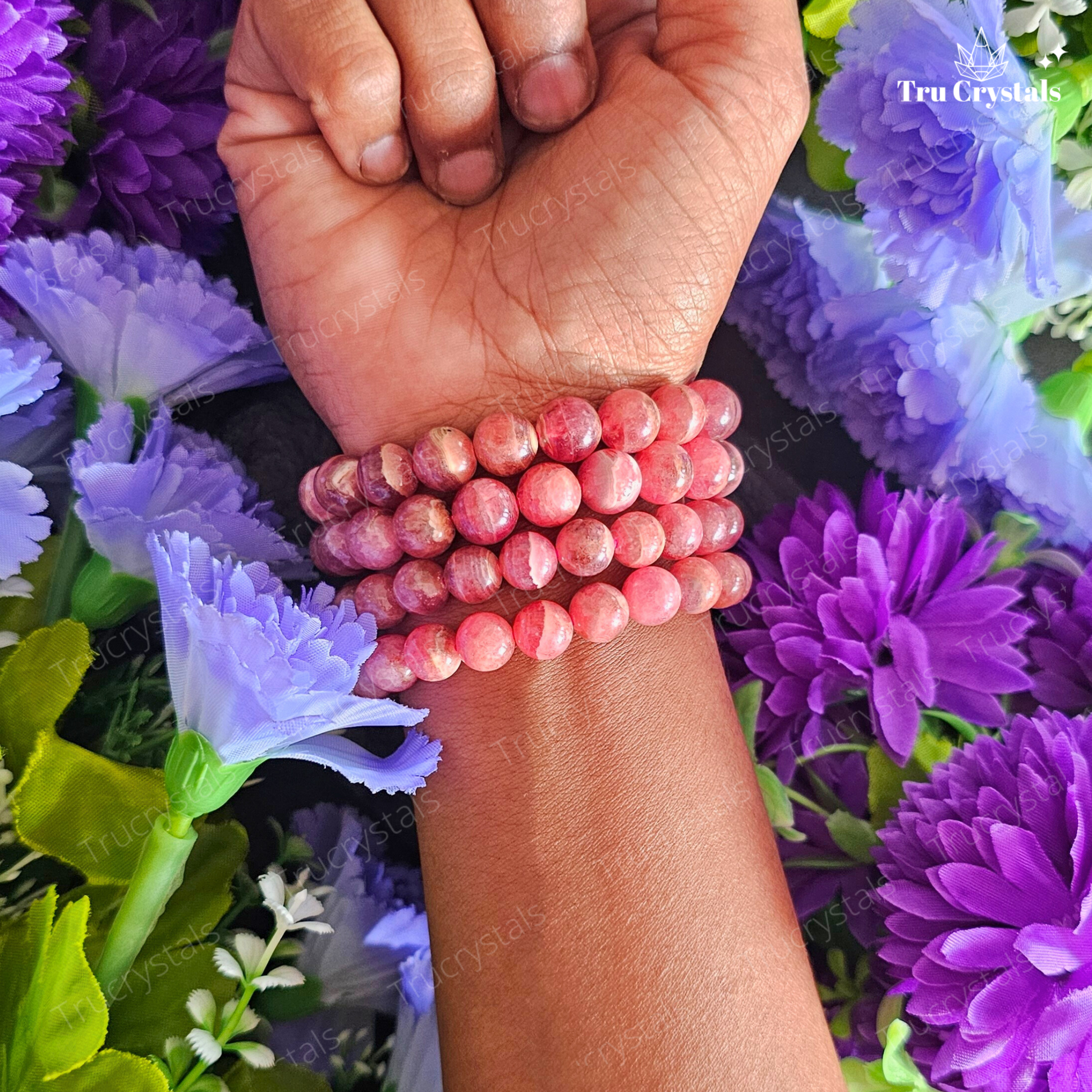A Rhodochrosite bracelet displayed on a dark stone surface with vibrant pink flowers around it. 