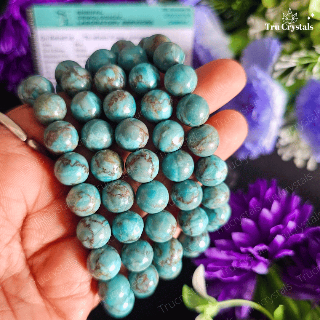 A South African Blue Turquoise 8mm & 10mm Bracelet displayed on a dark stone surface with vibrant purple flowers around it, including a certification card in the background.
