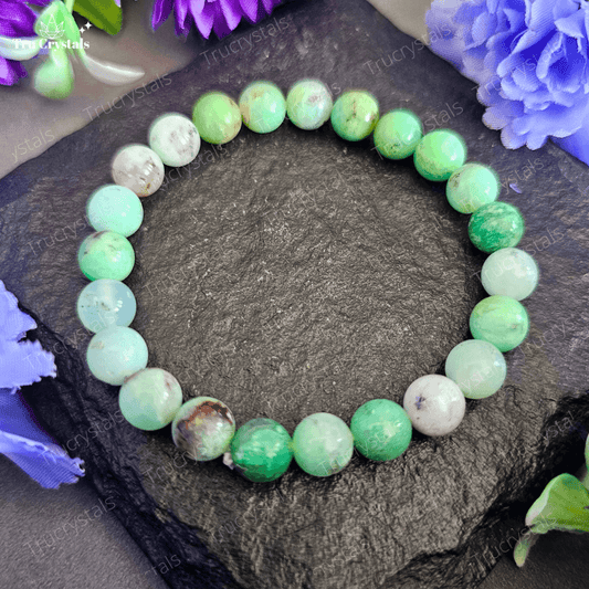 A bracelet featuring a series of round Chrysoprase gemstones on a stretch cord, displayed on a dark background with purple flowers around it.