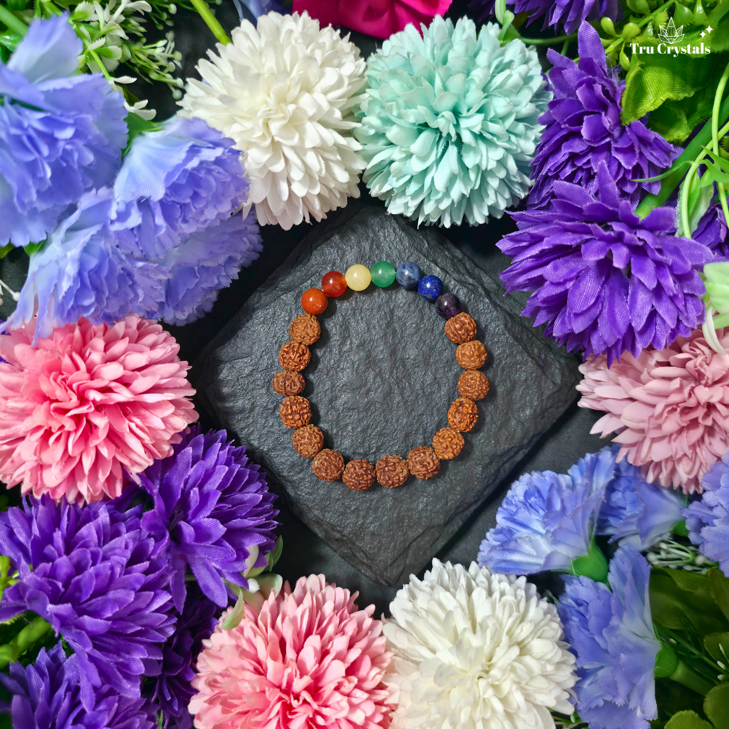 A beaded bracelet with brown rudraksha beads and a multi-colored chakra sequence, displayed on a dark slate square surrounded by colorful artificial flowers. The brand name 'TruCrystals' is visible in the top right corner.