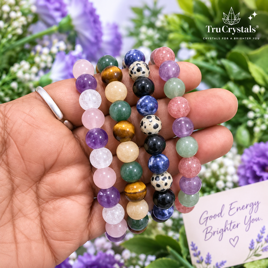 A hand holds four colorful beaded crystal bracelets, including amethyst, rose quartz, green aventurine, tiger's eye, lapis lazuli, and black-and-white spotted beads. The background is a blurred garden with purple flowers and green leaves, and the TruCrystals logo is visible in the top right corner.