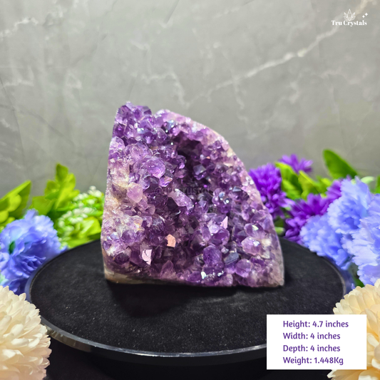 A natural amethyst crystal cluster geode specimen displayed on a black circular stand, surrounded by decorative artificial flowers against a gray textured wall. A white text box in the bottom right corner lists the dimensions and weight of the crystal.