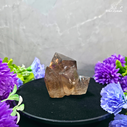 A natural smoky quartz crystal cluster displayed on a black circular stand, surrounded by purple and blue artificial flowers against a gray marble-patterned background. The brand name 'TruCrystals' is visible in the top right corner.