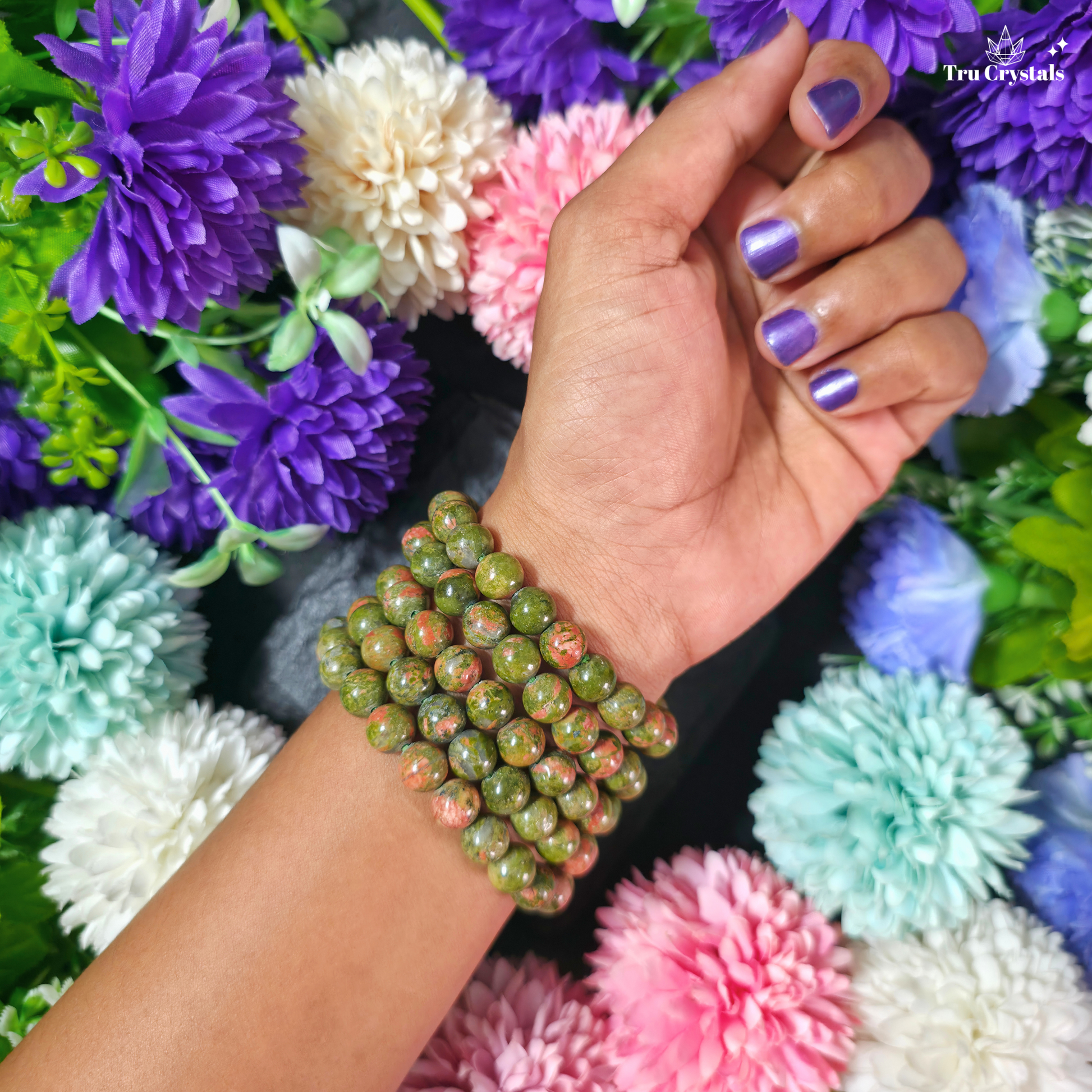 A close-up of a person's hand and wrist wearing a unakite beaded bracelet made of green and pink mottled stone beads, set against a background of colorful artificial flowers. The brand name 'True Crystal' is visible in the top right corner.