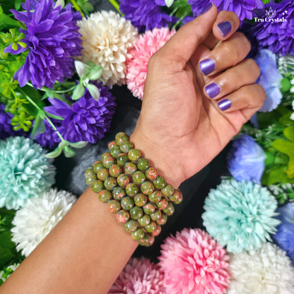 A close-up of a person's hand and wrist wearing a unakite beaded bracelet made of green and pink mottled stone beads, set against a background of colorful artificial flowers. The brand name 'True Crystal' is visible in the top right corner.