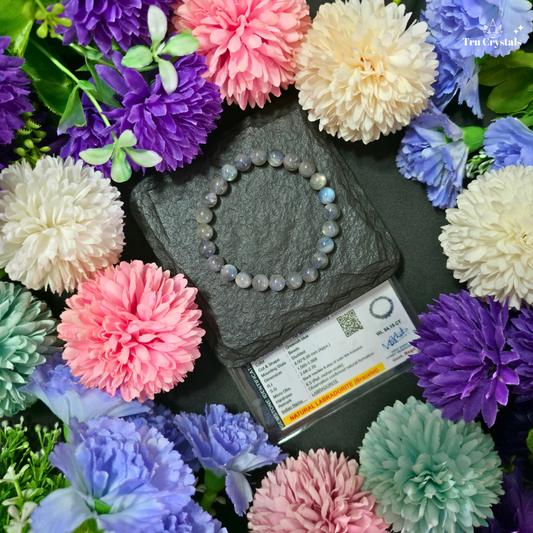 A Labradoritebeaded bracelet made of small round iridescent stones arranged in a circle on a dark slate block, surrounded by colorful artificial flowers and a small product card. The bracelet has a blue-gray base with shifting pastel highlights.