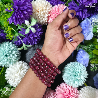 A close-up of a person's wrist wearing a multi-strand beaded bracelet made of deep red maroon beads, styled with colorful artificial flowers in purple, pink, white, and light blue. The brand name 'Tra Crystals' is visible in the top right corner.