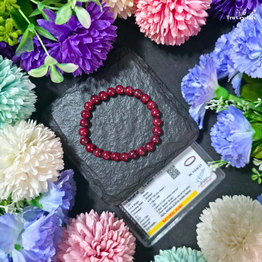 A dark natural red  ruby beaded bracelet arranged in a circle on a dark slate stone, surrounded by colorful artificial flowers and a product card. The brand name 'Trucrystals' is visible in the top right corner.