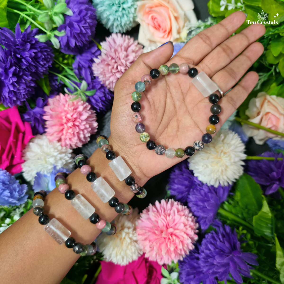 Three beaded stretch bracelets with clear rectangular crystal sections and dark round beads, displayed on a person's wrist and held in the hand against a background of colorful artificial flowers. The brand name 'Tru Crystals' is visible in the top right corner.