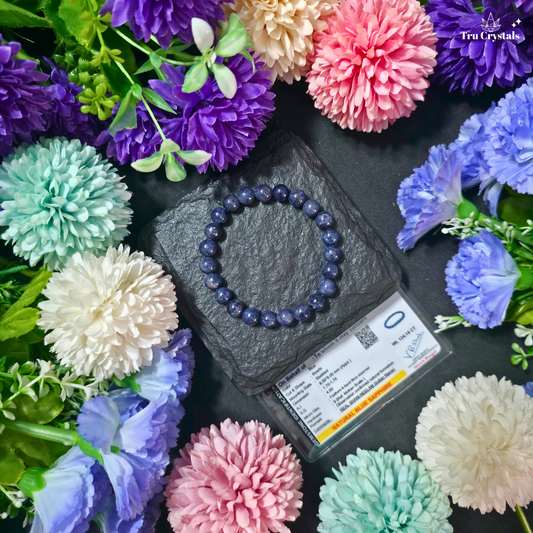 A beaded bracelet made with natural blue sapphire gemstones, displayed on a gray stone with purple and blue flowers around it carrying authentication card.