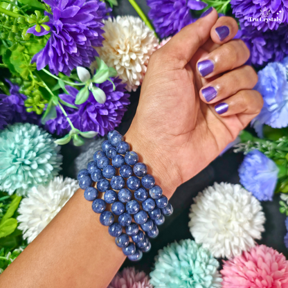 A close-up of a person's wrist wearing a blue sapphire beaded bracelet , set against a background of colorful artificial flowers.