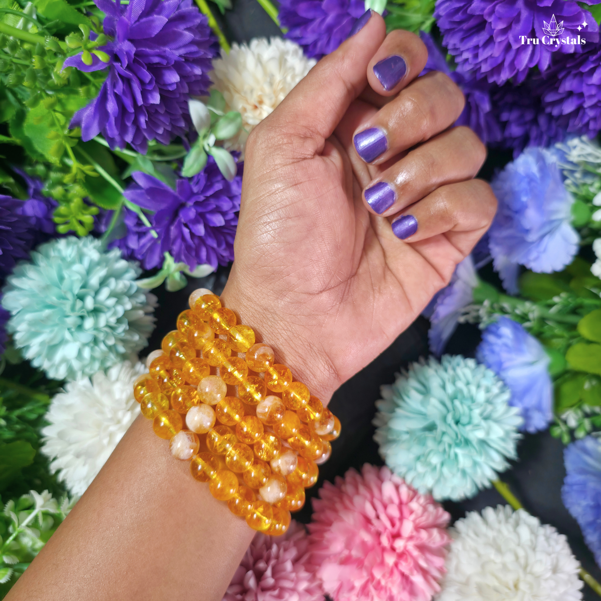 A close-up of a person's wrist wearing a stack of translucent amber-yellow beaded bracelets, with purple nail polish and a background of colorful artificial flowers. The brand name 'Ten Crystals' is visible in the top right corner.