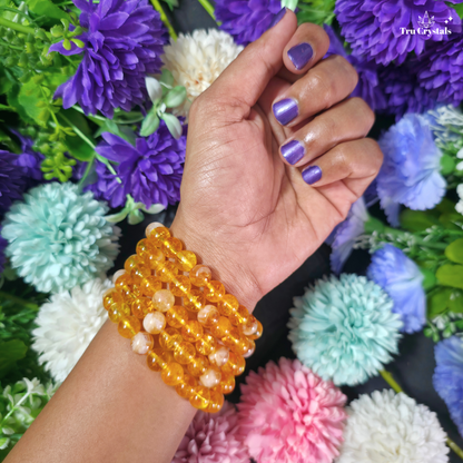 A close-up of a person's wrist wearing a stack of translucent amber-yellow beaded bracelets, with purple nail polish and a background of colorful artificial flowers. The brand name 'Ten Crystals' is visible in the top right corner.
