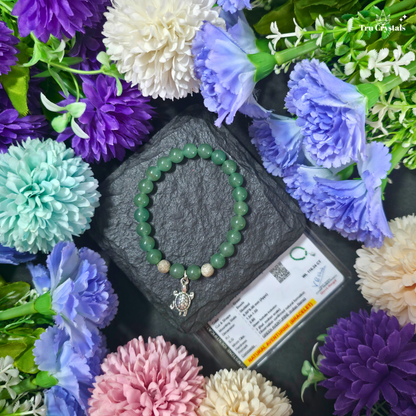 A green aventurine beaded bracelet with a turtle charm and two beige beads, displayed on a dark slate block surrounded by colorful artificial flowers. A product card is visible next to the bracelet.