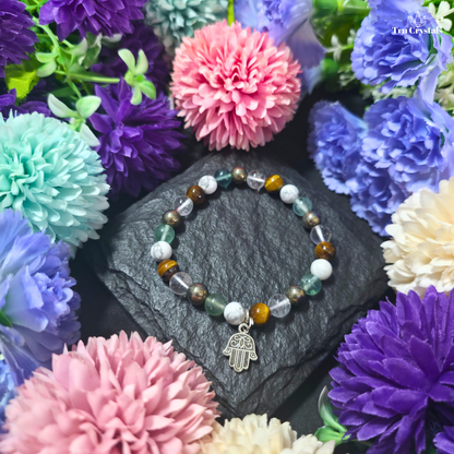 A multicolored student  bracelet featuring Tiger eye , fluorite , howlite,clear quartz and pyrite and a metal charm in the shape of a hand, displayed on a dark stone surface with flowers in the background.