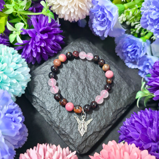 A multicolored stone  Soulmate bracelet displayed on a dark stone surface surrounded by various colorful flowers, with a small heart-shaped pendant hanging from the bracelet. The bracelet contains rose quartz , Rhodonite , rhodochrosite & garnet .