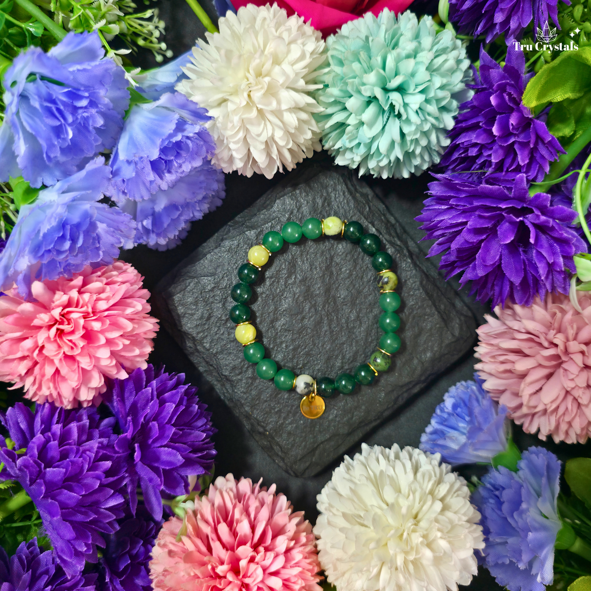 A green numerology bracelet with green jade, green aventurine, and serpentine stones, featuring a 'Be TRU' engraving on a black stone charm, displayed on a dark background with purple flowers.