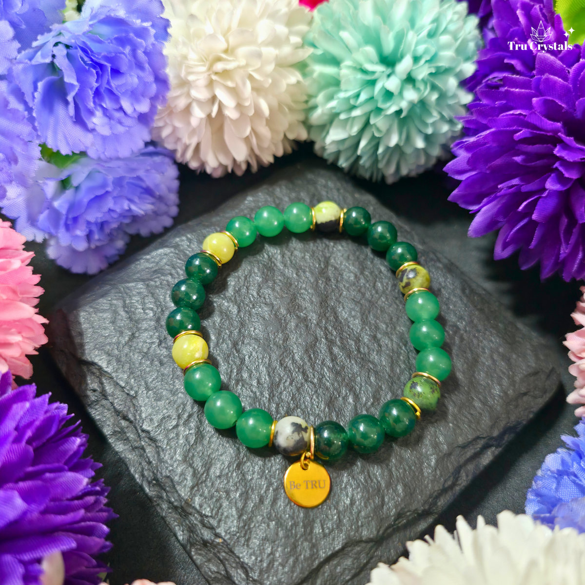 A green numerology bracelet with green jade, green aventurine, and serpentine stones, featuring a 'Be TRU' engraving on a black stone charm, displayed on a dark background with purple flowers.