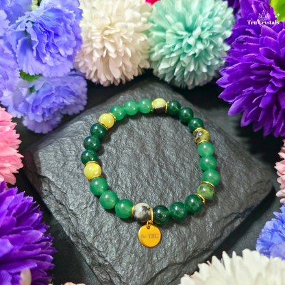 A green numerology bracelet with green jade, green aventurine, and serpentine stones, featuring a 'Be TRU' engraving on a black stone charm, displayed on a dark background with purple flowers.