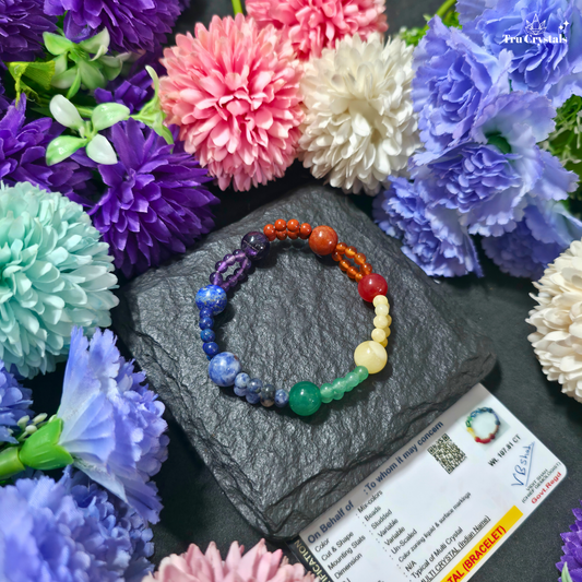 A multicolor beaded  7 chakra bracelet displayed on a dark gray stone slab, surrounded by colorful artificial flowers and a product card.