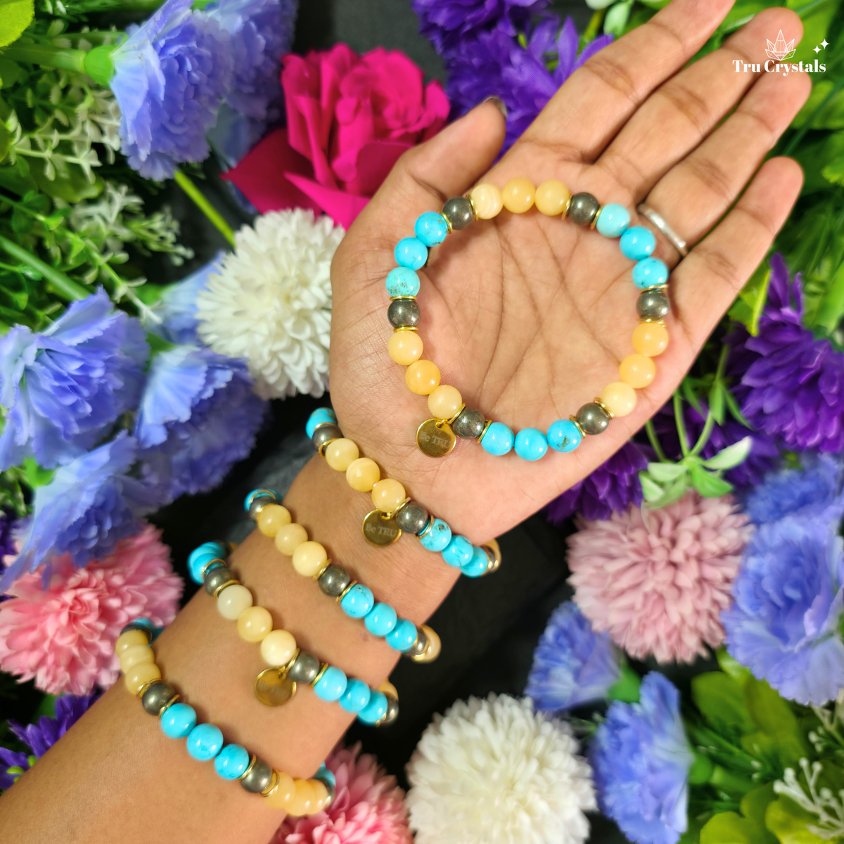 A hand holding three beaded bracelets, with two more stacked on the wrist, set against a background of colorful artificial flowers. The bracelets feature turquoise, yellow, and brown beads with small gold charms.