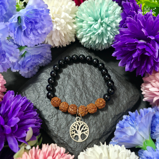 A black tourmaline beaded bracelet with Rudraksha seeds and a silver charm in the shape of the Tree of Life, displayed on a dark surface with flowers around it.