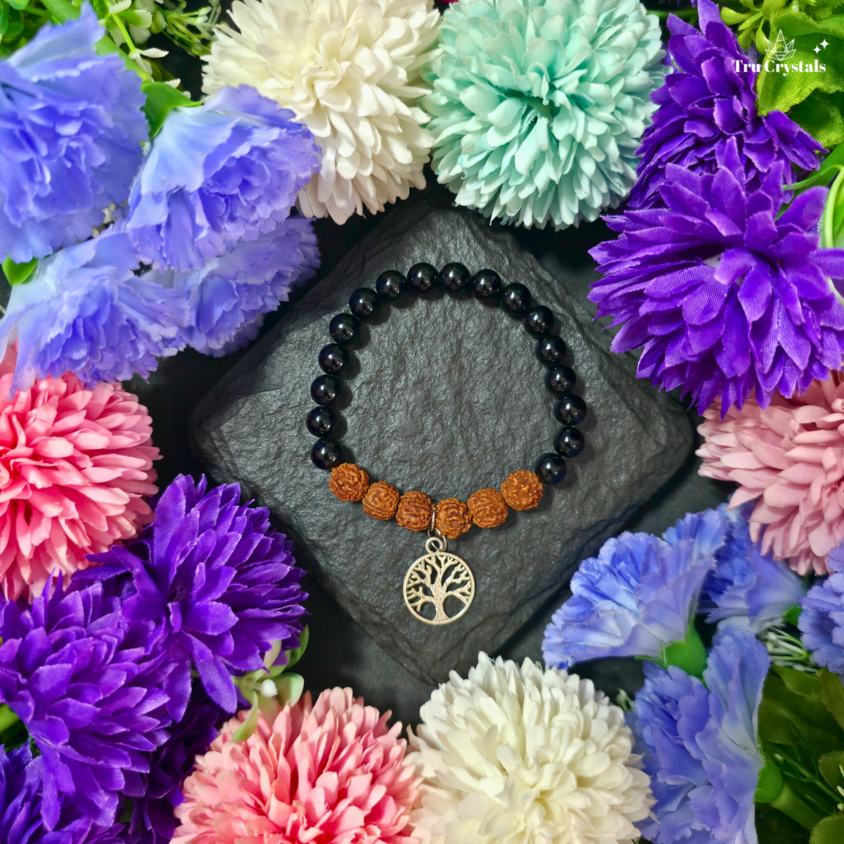 A beaded bracelet with black round beads, orange-brown textured beads, and a silver tree of life pendant, displayed on a dark stone surrounded by colorful artificial flowers. The brand name 'Trucrystals' is visible in the top right corner.