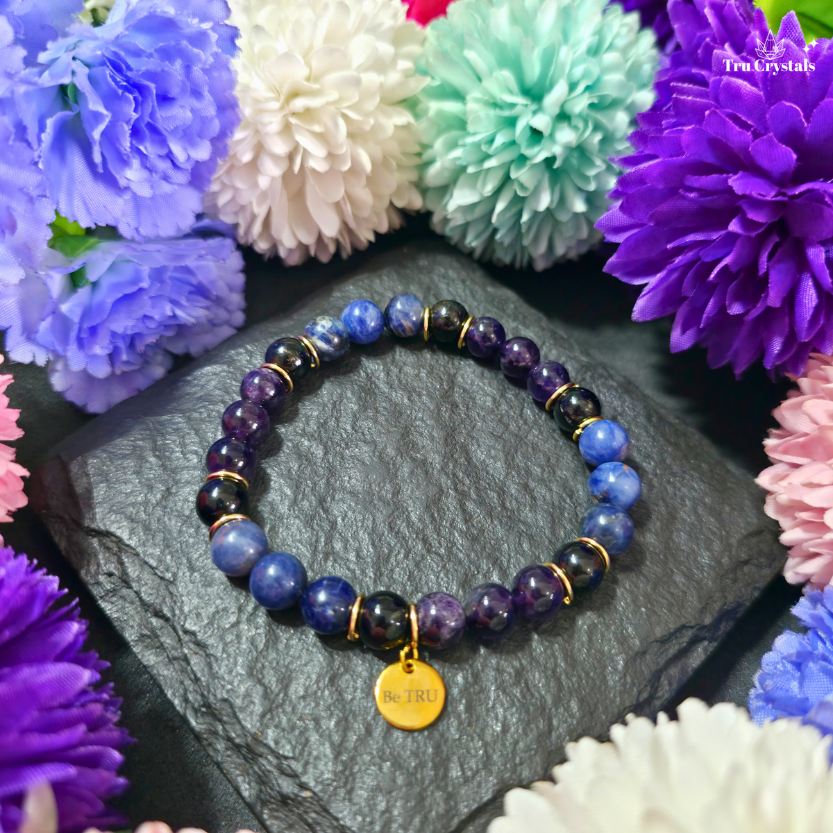 A bracelet with Sodalite, Amethyst, and Black Tourmaline  beads, featuring a 'Be TRU' engraved charm, displayed on a textured background with flowers.