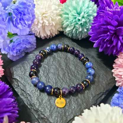 A bracelet with Sodalite, Amethyst, and Black Tourmaline  beads, featuring a 'Be TRU' engraved charm, displayed on a textured background with flowers.