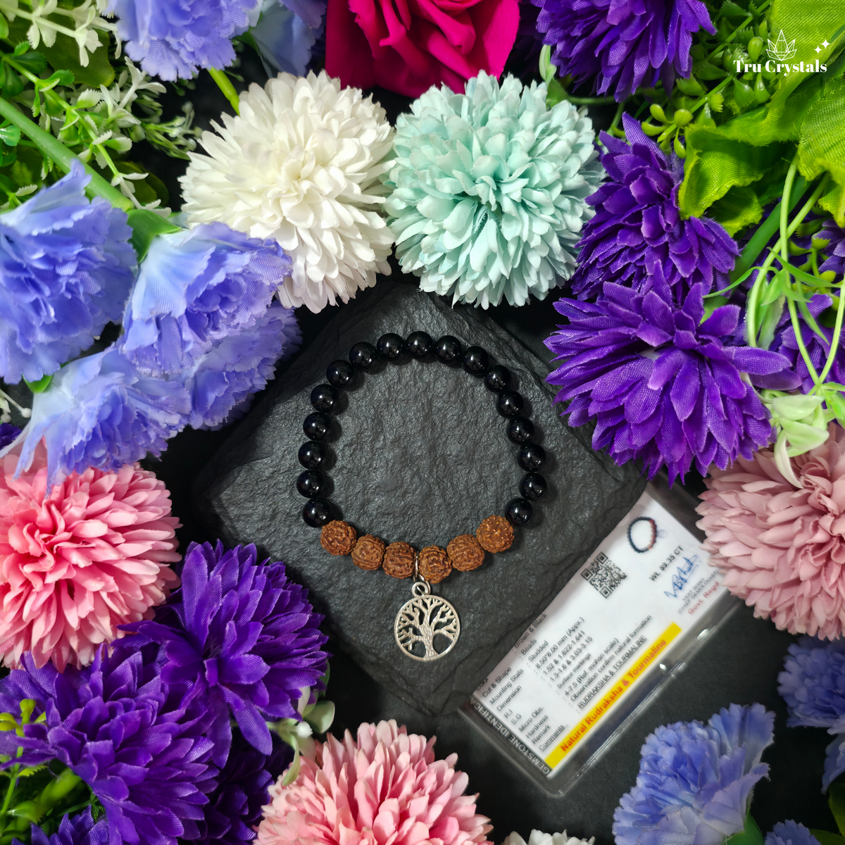 A beaded bracelet with black round beads, brown textured beads, and a silver tree of life pendant, displayed on a dark slate surrounded by colorful artificial flowers. A product card is visible next to the bracelet.
