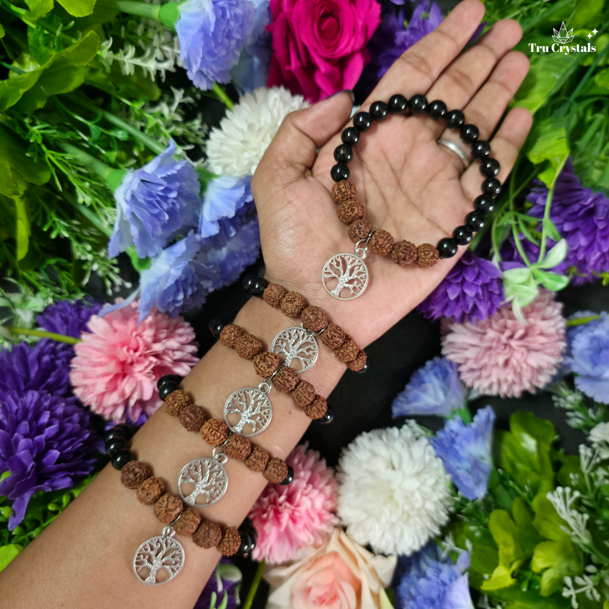 A hand and wrist wearing multiple beaded bracelets with brown rudraksha beads, black spacer beads, and silver tree-of-life pendants, posed against a background of colorful flowers. The brand name 'TruCrystals' is visible in the top right corner.
