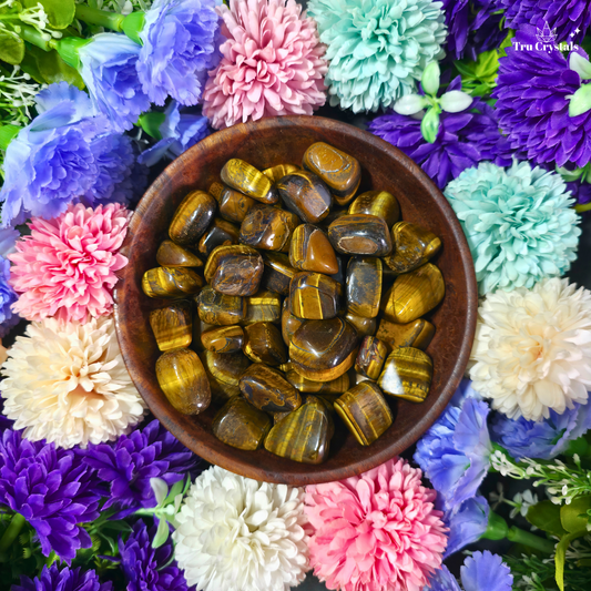 A top-down view of polished tiger's eye stones in a dark brown wooden bowl, surrounded by colorful artificial flowers including pink, purple, blue, and mint green.