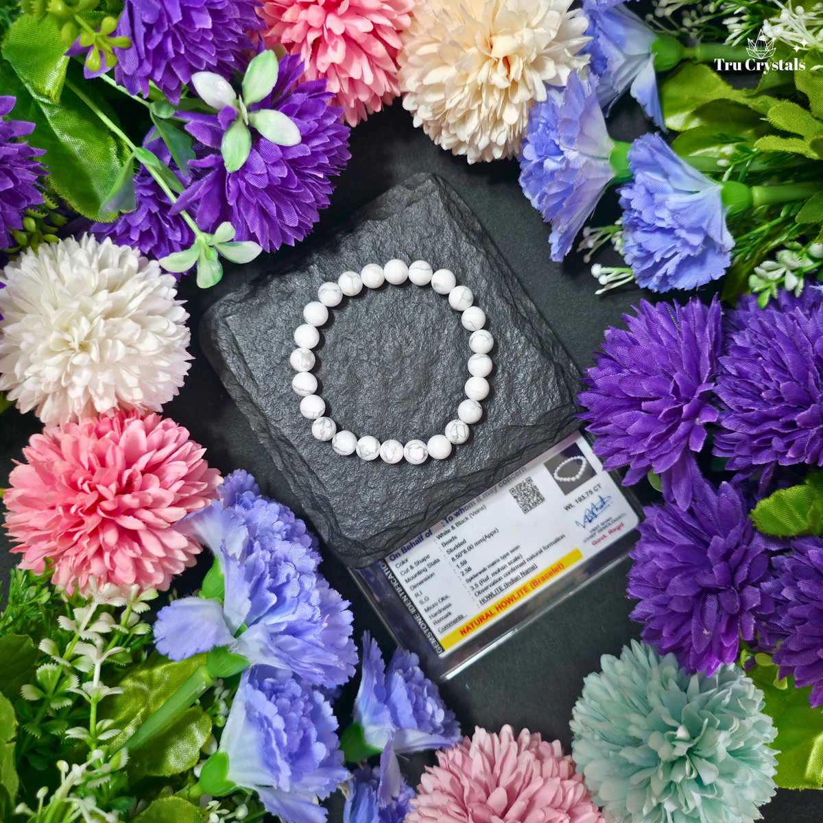 A white beaded  howlite bracelet arranged in a circle on a dark slate block, surrounded by colorful artificial flowers and a small branded card. The bracelet is the product, photographed in a styled flat-lay setup.
