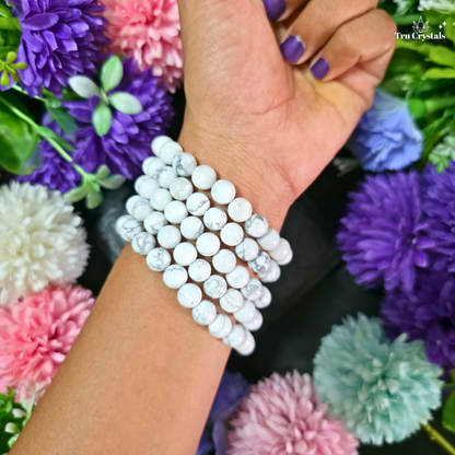 A close-up of a person's wrist wearing a stack of white howlite round beads, surrounded by colorful artificial flowers. The brand name 'TruCrystals' is visible in the top right corner.