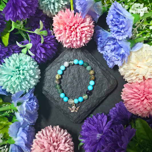 A multicolored beaded bracelet with a turquoise, moss agate , clear quartz and ocean jasper stone combination, displayed on a wooden surface with floral arrangements and crystal stones in the background.