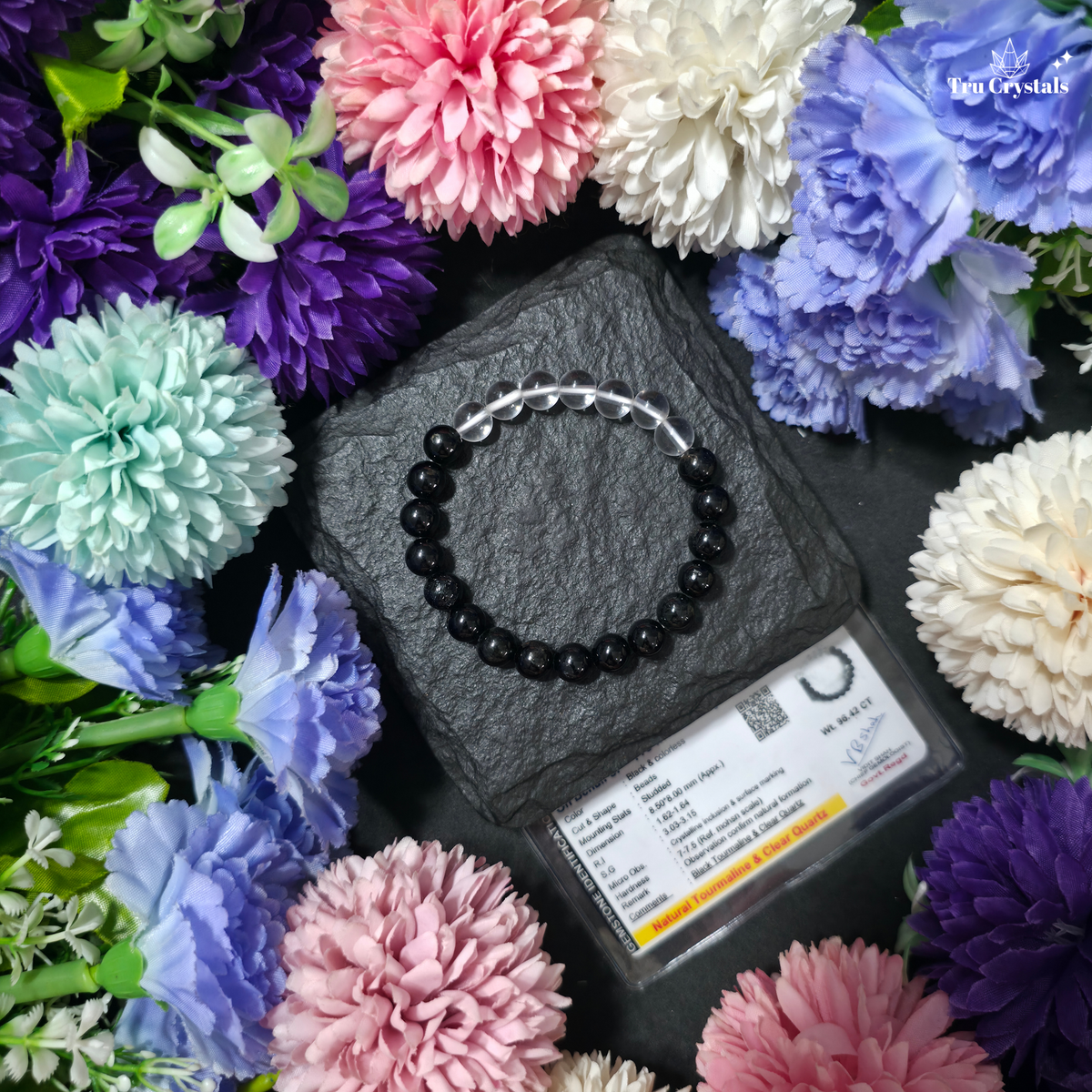 A multistone bracelet featuring black tourmaline and clear quartz, displayed on a textured background with certicate , colorful flowers and a dark stone in the foreground.